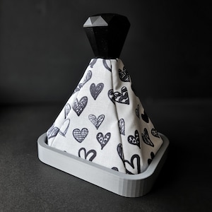 May include: A gray and white fabric gear shift knob with a black diamond-shaped top. The fabric has a pattern of black hearts on a white background.