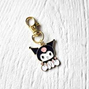 May include: A gold-coloured keychain featuring a black and white cartoon character with a skull on its head. The character has pink cheeks and is wearing a black dress with a white collar.