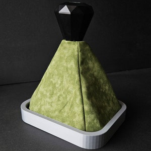 May include: A green fabric gear shift knob with a black diamond-shaped top. The knob is mounted on a white base.