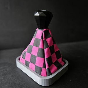 Peut inclure: Un pommeau de levier de vitesse à damier noir et rose avec un dessus en forme de diamant noir. Le pommeau est monté sur une base grise.