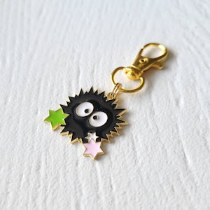 May include: A gold keychain featuring a black and white cartoon character with large eyes and green and pink stars.