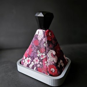 May include: A decorative bottle stopper featuring a black, geometric top and a floral fabric skirt in purple, pink, and red hues. The stopper is set in a grey, triangular base.