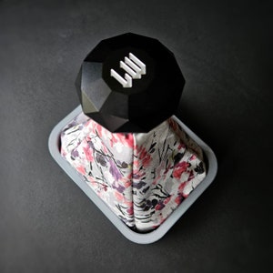 May include: A black, geometric-shaped object with white text on top of a grey, square-shaped container. The container holds a floral patterned fabric with pink and purple flowers.