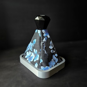 May include: A black and blue gear shift knob cover with a floral pattern. The cover is made of fabric and has a diamond-shaped top. The cover is attached to a grey base.