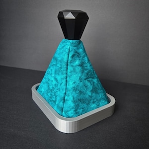 May include: A gear shift boot featuring a textured teal fabric cover, a black faceted diamond-shaped knob at the top, and a rectangular light gray base. The assembly is designed for automotive interior use with a distinct geometric aesthetic.