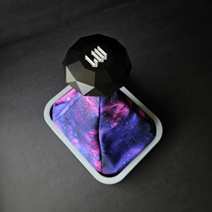 May include: A black, geometric gear shift knob with the letters "LW" on top, sits on a purple and pink galaxy print gear shift boot.