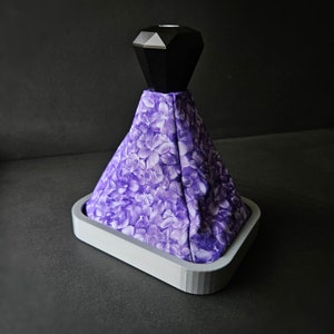 Puede incluir: Cubierta de pomo de palanca de cambios de tela floral morada con una parte superior negra en forma de diamante. La cubierta está unida a una base gris.