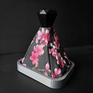 May include: A black and pink floral patterned gear shift knob with a black diamond-shaped top. The knob is mounted on a grey base.