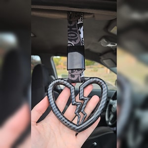 May include: A car grab handle featuring a broken heart design. The handle is made of a carbon fiber material with a black and gray pattern. The strap is black and white with a pattern. The handle is held in a person's hand.