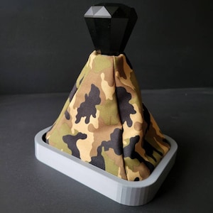 May include: A custom car shift boot with a black faceted knob and a camouflage fabric cover. The boot is set in a gray rectangular base, offering a unique and stylish interior upgrade.
