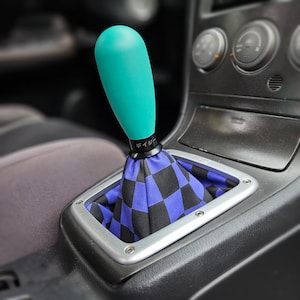 May include: A teal shift knob with a black base and Japanese text. The shift boot has a black and blue chequered pattern. The assembly is set in a silver frame within a car's console.