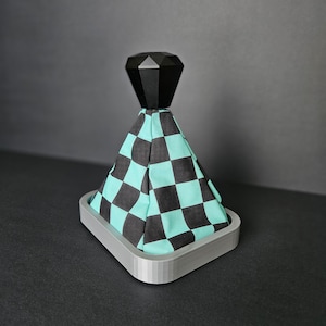 Peut inclure: Un pommeau de levier de vitesses à damier noir et turquoise avec un dessus en forme de diamant noir. Le pommeau est monté sur une base grise imprimée en 3D.