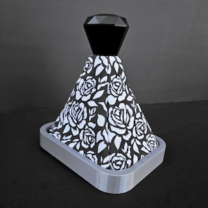 May include: A black and white floral patterned gear shift knob with a black diamond-shaped top. The knob is mounted on a grey base.