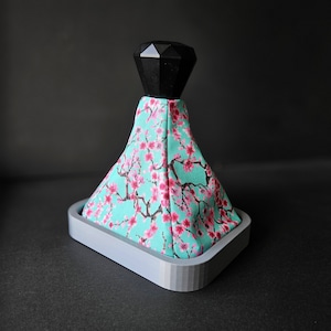 May include: A grey, square-shaped base with a black diamond-shaped knob on top. The base is covered in a turquoise fabric with a pink cherry blossom pattern.