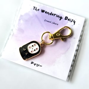 May include: A gold-coloured keychain featuring a black and white enamel charm of No Face from the film Spirited Away. The charm is attached to a gold-coloured metal ring and a gold-coloured lobster clasp. The keychain is packaged on a white card with the text "The Wandering Daisy Enamel Charm" and "デイジー" in Japanese.