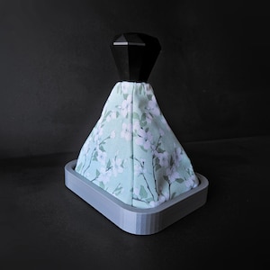 May include: A grey, square-shaped gear shift knob with a black diamond-shaped top. The knob is covered in a light green fabric with a white floral pattern.