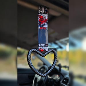 May include: A car grab handle with a heart-shaped grip. The strap features a Spider-Man comic book design in red, black, and white. The grip has a carbon fiber pattern.