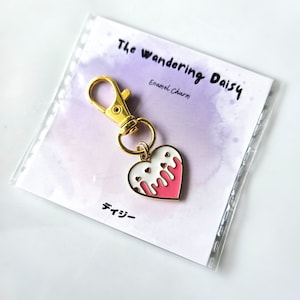 May include: A gold-tone metal charm with a lobster clasp, featuring a pink heart with white dripping accents and small red hearts. The charm is attached to a card with the text "The Wandering Daisy" and "Enamel Charm".