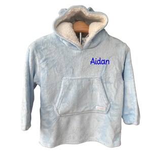 May include: A light blue hooded blanket with a large front pocket and the name "Aidan" embroidered in blue. The hood has small bear ears and a white, fluffy interior. The blanket is made of a soft, plush material.