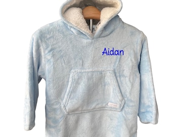 Puede incluir: Una manta con capucha azul claro con un bolsillo frontal grande y el nombre "Aidan" bordado en azul. La capucha tiene pequeñas orejas de oso y un interior blanco y esponjoso. La manta está hecha de un material suave y afelpado.