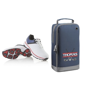 May include: White golf shoes with red and blue accents and a navy blue golf shoe bag with a gray zipper and a white embroidered name "THOMAS" with crossed golf clubs and a golf ball.