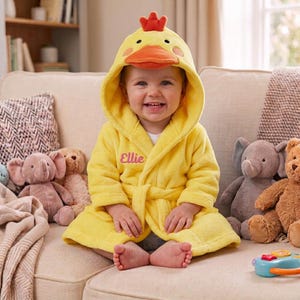 May include: A bright yellow hooded bathrobe featuring a duck design with an orange beak and a red crest on the hood. The robe has long sleeves, a tie belt, and the name Ellie embroidered in pink script on the left side of the chest area.