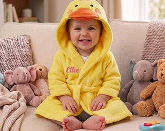 Puede incluir: Una bata de baño con capucha de color amarillo brillante con diseño de pato, que incluye un pico naranja y una cresta roja en la capucha. La bata tiene mangas largas, un cinturón y el nombre Ellie bordado en rosa en el pecho izquierdo.
