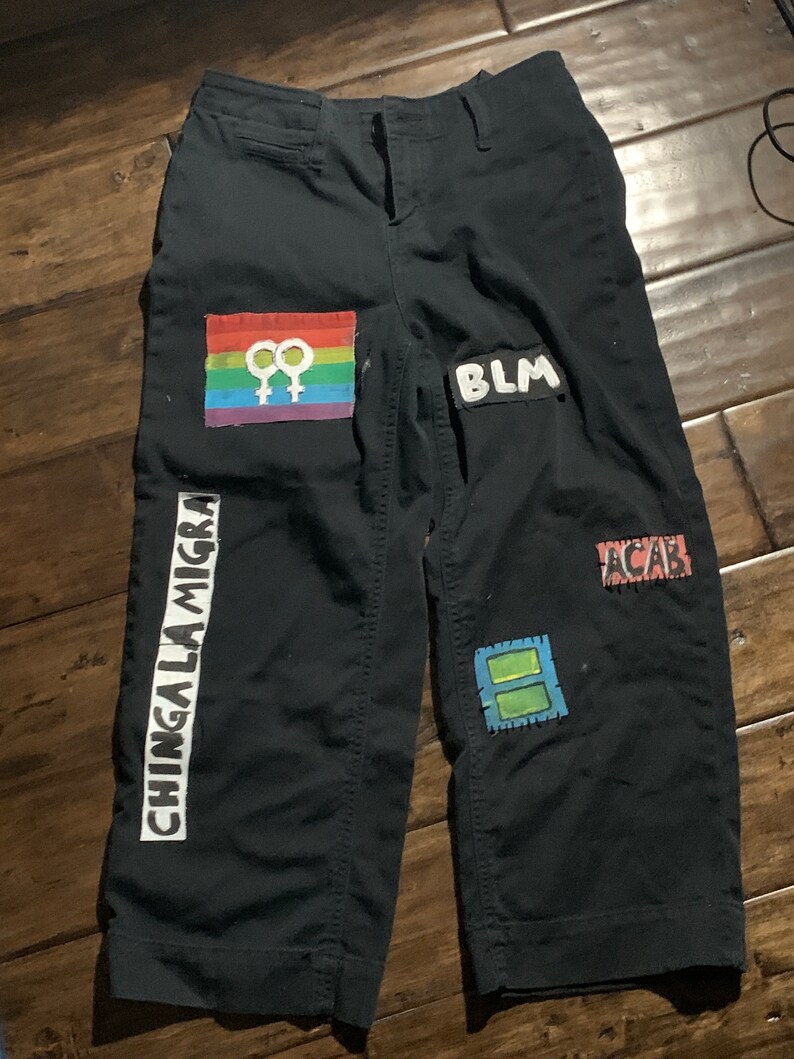 K&ouml;nnte beinhalten: Schwarze Hose mit verschiedenen aufgen&auml;hten Patches. Die Patches beinhalten eine Regenbogenflagge mit dem weiblichen Symbol, die Buchstaben "BLM", die Buchstaben "ACAB" und die Worte "CHINGA LA MIGR",