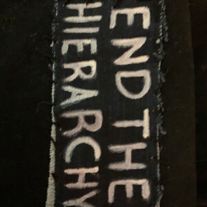 K&ouml;nnte beinhalten: Ein schwarzer Stoff-Patch mit wei&szlig;em Text, der "END THE HIERARCHY" lautet. Der Patch hat ausgefranste Kanten und ist auf einen schwarzen Hintergrund gen&auml;ht.