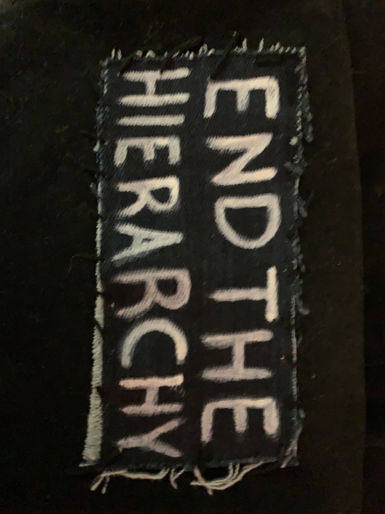 K&ouml;nnte beinhalten: Ein schwarzer Stoff-Patch mit wei&szlig;em Text, der "END THE HIERARCHY" lautet. Der Patch hat ausgefranste Kanten und ist auf einen schwarzen Hintergrund gen&auml;ht.