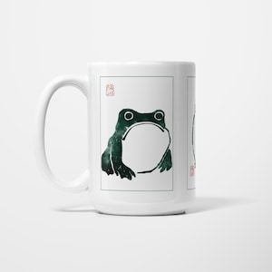 May include: White ceramic mug with a dark green frog illustration. The frog has a large open mouth and is framed within a rectangular border. A red stamp is in the upper left corner.