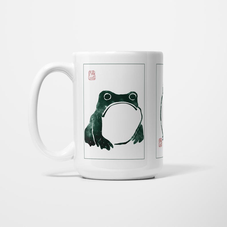 May include: White ceramic mug with a dark green frog illustration. The frog has a large open mouth and is framed within a rectangular border. A red stamp is in the upper left corner.