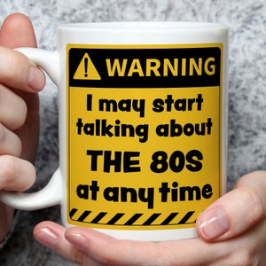 Puede incluir: Taza de cer&aacute;mica blanca con una se&ntilde;al de advertencia amarilla y negra que dice "ADVERTENCIA Puedo empezar a hablar de los 80 en cualquier momento".