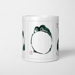 May include: White ceramic mug with a dark green ink illustration of a frog. The frog is depicted in a minimalist style with a circular body and two eyes. The mug has a red stamp on the side.