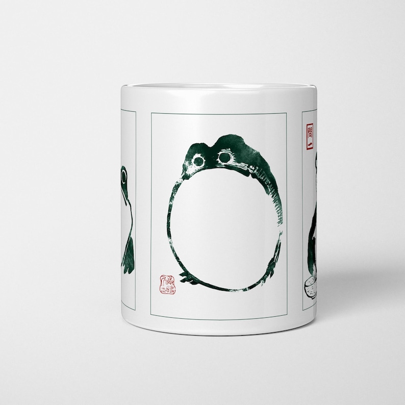 May include: White ceramic mug with a dark green ink illustration of a frog. The frog is depicted in a minimalist style with a circular body and two eyes. The mug has a red stamp on the side.