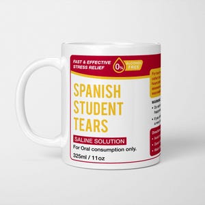 May include: A white ceramic mug featuring a red and yellow label that reads "SPANISH STUDENT TEARS" in large yellow text. The label includes "FAST & EFFECTIVE STRESS RELIEF," "SALINE SOLUTION," "For Oral consumption only," and "325ml / 11oz."