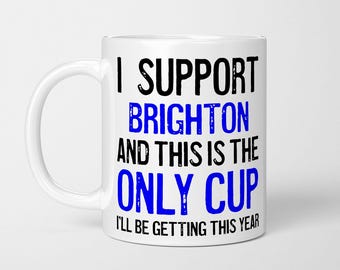 May include: A white ceramic mug featuring black and blue text that reads: I SUPPORT BRIGHTON AND THIS IS THE ONLY CUP I'LL BE GETTING THIS YEAR. The mug has a standard handle on the left side and a smooth, cylindrical body with a glossy finish.