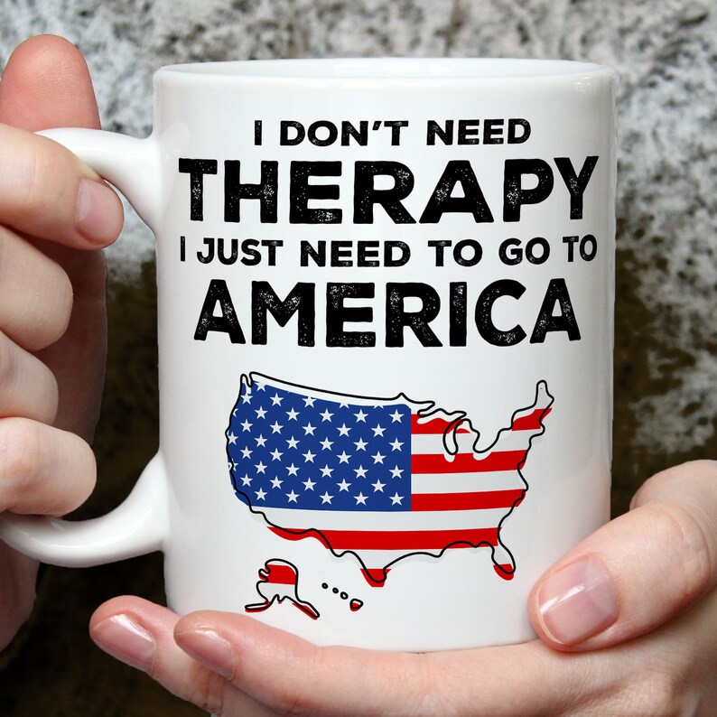 K&ouml;nnte beinhalten: Wei&szlig;e Keramiktasse mit einer schwarz-wei&szlig;en Grafik, auf der "Ich brauche keine Therapie, ich muss nur nach Amerika" steht, und einer rot-wei&szlig;-blauen Umrandung der Vereinigten Staaten.