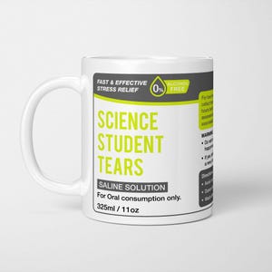 May include: A white ceramic mug featuring a label with the text Science Student Tears, Saline Solution, 325ml / 11oz, and Fast & Effective Stress Relief. The design includes lime green and dark grey accents, with additional warning and direction text.