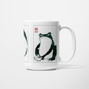 May include: White ceramic mug with a Japanese-style frog illustration. The frog is drawn in dark green ink, holding a bowl, and has a stern expression. A red Japanese character is also present on the mug.