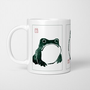 May include: White ceramic mug with a dark green frog design. The frog illustration has large eyes and a wide mouth, with a Japanese-style stamp. The mug is a standard size for tea or coffee.