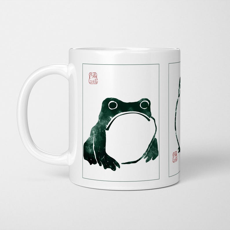 May include: White ceramic mug with a dark green frog design. The frog illustration has large eyes and a wide mouth, with a Japanese-style stamp. The mug is a standard size for tea or coffee.