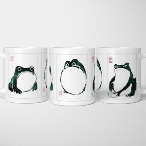 May include: White ceramic mugs featuring a black and green frog illustration in a Japanese ink wash style. Each mug has a handle on the right. The frog art is on the front of the mug.