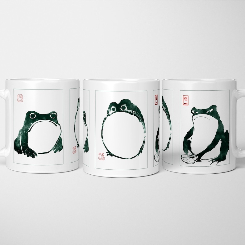 May include: White ceramic mugs featuring a black and green frog illustration in a Japanese ink wash style. Each mug has a handle on the right. The frog art is on the front of the mug.