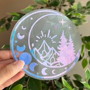 May include: A round, iridescent sticker featuring a mountain range, pine trees, crescent moons, and stars. The design is set within a larger crescent moon shape. The sticker has a holographic effect, reflecting various colors.