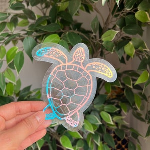 Könnte beinhalten: Ein transparenter, schimmernder Aufkleber mit einer Meeresschildkröte in einem Farbverlauf von Pink, Blau und Gelb. Der Aufkleber ist mit einem schwarzen Umriss versehen.
