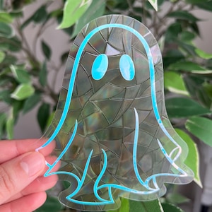 May include: A clear, ghost-shaped sticker with a holographic, textured background. The ghost design features a light blue outline and matching oval eyes. The sticker is held in front of a background of green leaves.