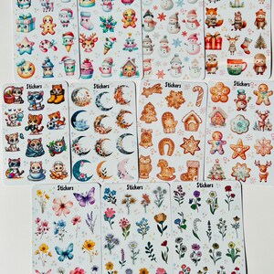 May include: A collection of sticker sheets featuring various designs. The sheets include stickers with winter holiday themes like snowmen and gingerbread men, along with floral and celestial designs. Each sheet is labeled with the word "Stickers".