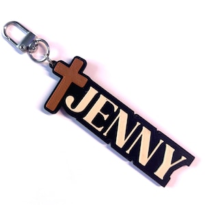 Peut inclure: Un porte-clés avec le nom "JENNY" en lettres crème sur fond noir, avec une croix marron. Le porte-clés est équipé d'un fermoir argenté.