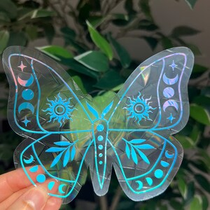 May include: A clear iridescent sticker of a butterfly with blue and white celestial designs, including moons, stars, and suns.
